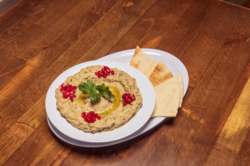 Baba Ghanoush with pomegranate seeds, parsley, and olive oil, served with pita bread on a wooden table.